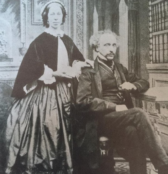 A black and white photograph of John Fisher and his wife. John Fisher is sat at his desk. His wife stands beside him. The room they are in is richly furnished, with large windows. A grand fireplace can be seen behind the couple.