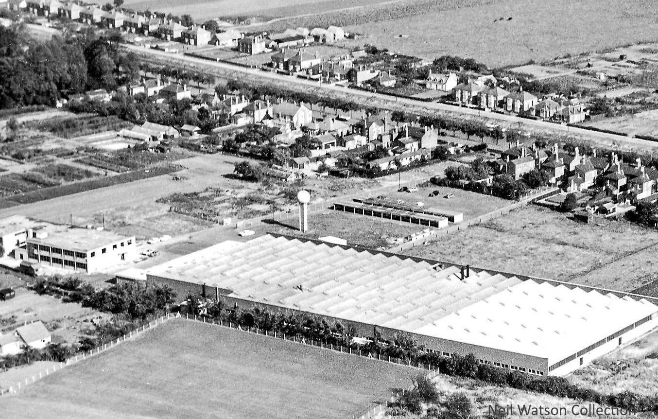 A black and white birds eye photograph of the Norprint factory. It is a large building with a long row of windows across the side. There are houses, allotments and scrub land surrounding the factory.