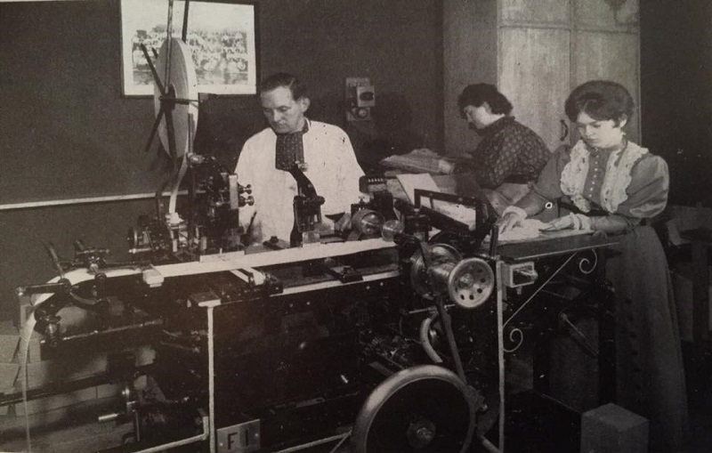 A black and white photograph of a printing machine. It is operated by a man and two women. The man is dressed in a white coat, while the women wear floor length dresses. The printing machine has a long strip of white fabric fed through it, and there are multiple wheels and belts across its construction.