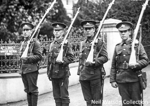 A black and white photograph of four soldiers stood in uniform, holding their guns at rest on their left shoulder. They are looking directly at the camera. The soldiers are Men of the 4th Lincolnshires