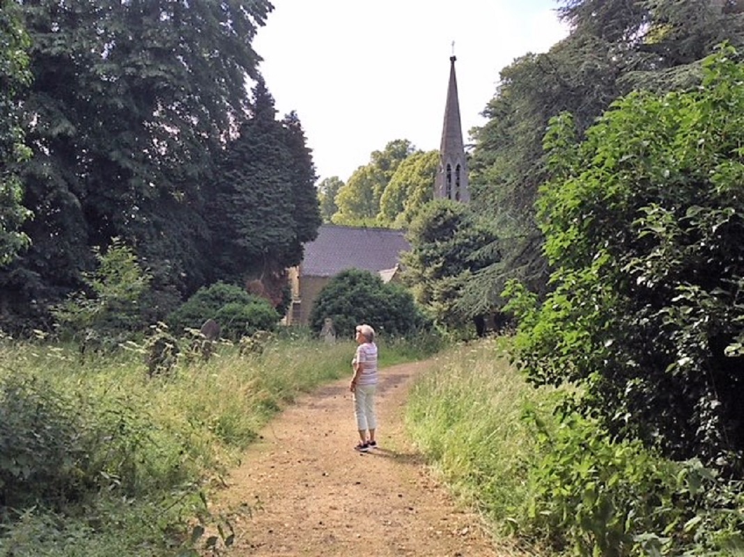 A coloured photo of a woman bird spotting on a sunny day. The woman is stood on a country path, with a church and spire in the background.