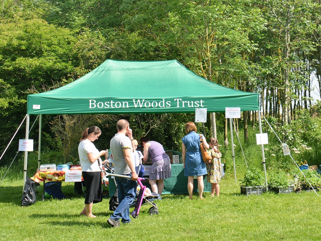 A photograpg of Boston Woods Trust's Open Day Sale. There is a green tent pitched in a wooded area. Several people are gathered around looking at items for sale, including cakes.